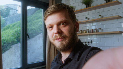 Bearded man looks directly at camera with a gentle smile, suggesting a friendly moment during a video call in natural light.