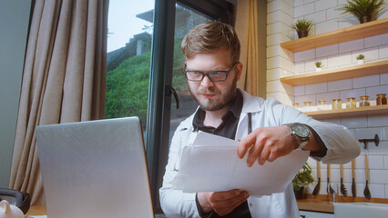 Male doctor wearing glasses and a lab coat reviews printed medical documents while working on a...
