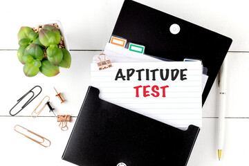 Aptitude test text on lined index card in black folder with office supplies and green plant on white wooden desk