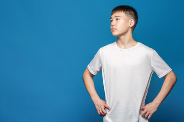Portrait of young handsome asian boy in white T-shirt holding hands on his belt against blue background.
