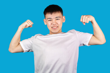 Portrait of handsome smiling Asian young boy in white t-shirt raising clenched fists and tensing muscles against blue background. Teen boy showing strength and power