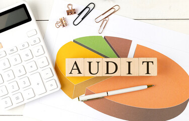 Audit word on wooden blocks placed over financial pie chart with calculator, pen, paper clips