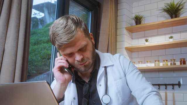 Bearded doctor in white coat speaks on phone with a serious expression while working at a laptop near a window in office setting.