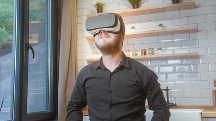 Bearded man in black shirt uses a VR headset in a bright modern kitchen, enjoying immersive digital content or virtual exploration.
