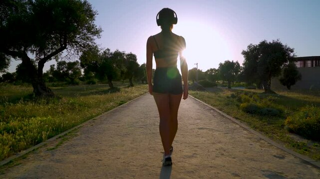 Mujer joven deportista caminando y escuchando m&uacute;sica por el parque al atardecer con el sol en el fondo a contraluz.