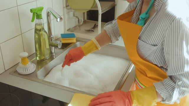 A soapy sink hints at a clogged drain. Woman stirs the water, facing a common plumbing issue. Cleaning cloth and spray bottle nearby, seeking a solution.