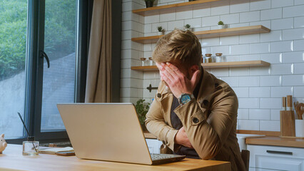 Man in beige jacket sits at desk, covering his face in stress while using a laptop, indicating...