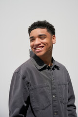 Obraz premium Young Dominican man with a bellicut or taper fade smiles widely as he leans casually against a plain white wall. He wears a dark gray shirt and projects a relaxed and cheerful mood.