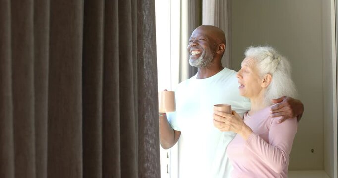 Sunlight spilling through drapes drawing diverse senior couple sipping coffee and laughing at home