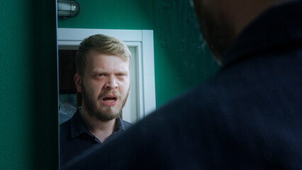 Fototapeta premium Troubled man appears to argue with his reflection in bathroom mirror, suggesting internal conflict, mental struggle, or emotional crisis.