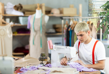 Professional young attentive man seamstress sewing textile using sewing machine sitting at table at...