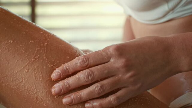 A young woman applies a body scrub while standing in a shower. Soft light filters through the window, creating a relaxing atmosphere for her beauty routine. She focuses on skin care.