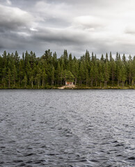 Hikers hut (Slogbod) hidden behind a pine tree along the shore of a serene lake in Jämtland Län dense forests, Sweden, creating a peaceful natural retreat with overcast sky