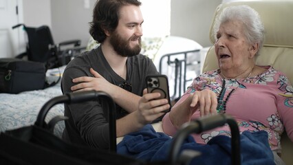 Fototapeta premium A grandson shares joyful moments with his grandmother while showing pictures on a smartphone. They enjoy smiles and laughter during this heartwarming visit.