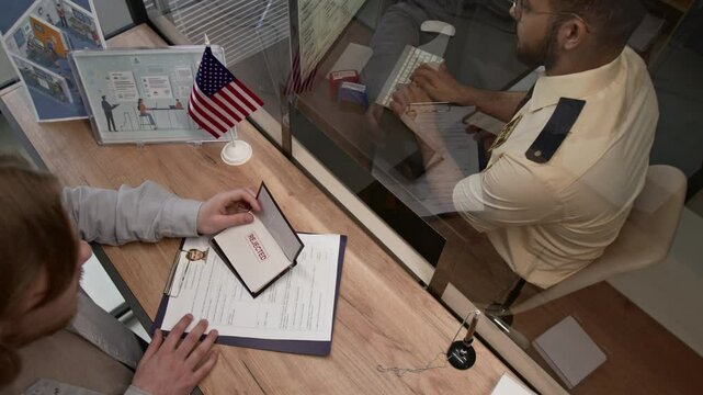 View from above of male submission officer returning rejected visa application to frustrated young man and proceeding to work on computer