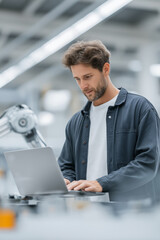 Engineer works on laptop in modern facility with robotic arm assisting in automation tasks