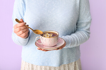 Beautiful young woman with cup of chamomile tea near lilac wall