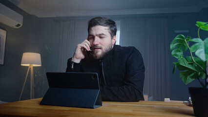 A businessman in a dark sweater is seated at a desk at home, engaged in a phone call while working on tablet. Quiet and focused ambiance for remote work.