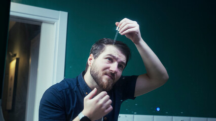 A man in the bathroom applying hair growth serum to his scalp. Massages the product into his hair while standing in front of the mirror.
