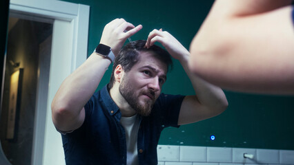 Man in the bathroom applying hair growth serum to his scalp. Massages the product into his hair while standing in front of the mirror