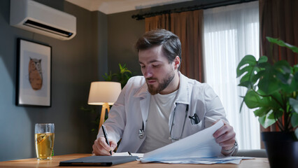 A doctor wearing a white coat carefully reviews a document while taking notes in a notebook, showing focus and professionalism in his medical work