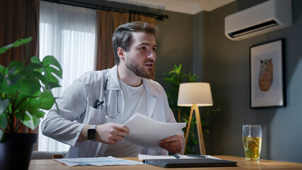 Male doctor wearing glasses and a stethoscope reviews patient documents in a modern office. With a focused expression, he analyzes medical records