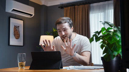 Young man in a casual white shirt participates in an online video call meeting at home. He holds up paper reports to show to the camera while actively talking and explaining.