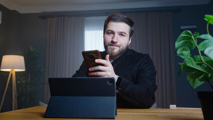 Man sits in a modern room holding smartphone in one hand while looking at a tablet on the desk. Focused man reviewing documents or working on administrative tasks