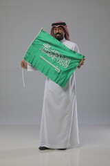 
Saudi man proudly holding the Saudi Arabian flag while smiling, wearing traditional dress. Studio shot expressing patriotism and national pride.