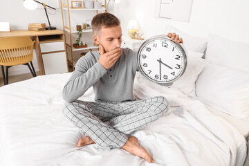 Young man holding clock and sitting on bed at home. Insomnia concept