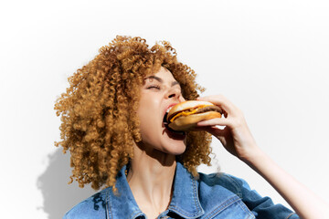 Young woman with curly hair joyfully eating a burger against a minimalist white background, showcasing vibrant colors and a casual style for healthy eating concepts