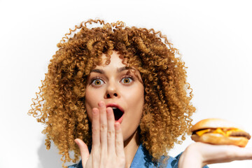 Excited young woman with curly hair, holding a burger, expressing surprise and joy, perfect for healthy eating concepts and seasonal promotions