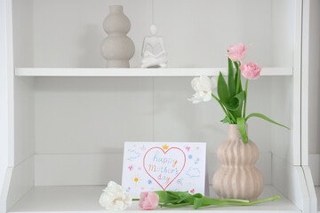 Vase with flowers and card for Mother's Day on shelf in room