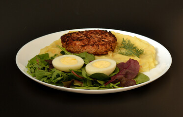 Plate with tasty lunch, meat cutlet on mashed potatoes, halves of boiled chicken egg and greens on black background.