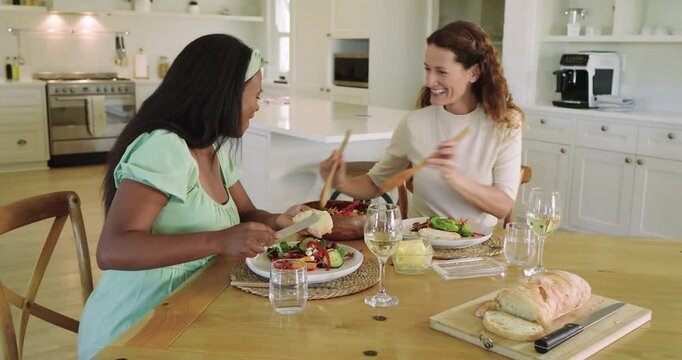 Diverse female friends upon sitting buttering baguette and tossing salad in kitchen chatting