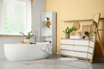 Interior of stylish bathroom with bathtub, shelving unit, mirror and green blossoming branches in...