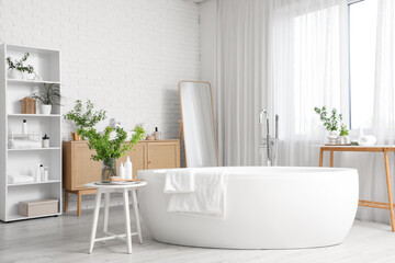 Interior of stylish bathroom with bathtub, shelving unit, mirror, commode and green blossoming branches in vases on tables
