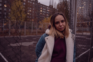 A young woman in a cozy jacket leans against a chain link fence at an urban park during autumn in...