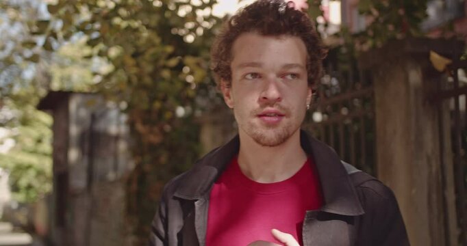 A young man with curly hair and a goatee walks down a sunny street while looking around and talking. He wears a red shirt and a black jacket.