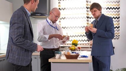 Three men in business clothes are standing in the office kitchen and eating sandwiches. Lunch break at work
