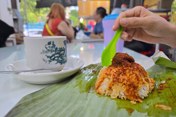  Close-up of a nasi lemak wrapped in banana leaf with sambal and meat, served alongside a vintage kopitiam-style coffee cup on a table in a local eatery in Malaysia.