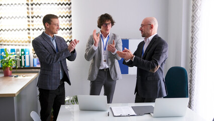Three successful, confident men in suits are standing around a table with laptops and applauding each other. Businessmen congratulate each other