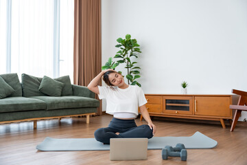 Young Asian woman stretching yoga workout on exercise mat while online training class with computer laptop in living room