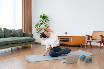 Young Asian woman stretching yoga workout on exercise mat while online training class with computer laptop in living room