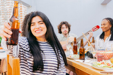 Young latin toasting with beer and smiling at the camera, her friends sing and play guitars