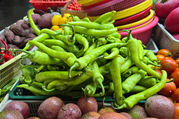 A close-up of vibrant green chili peppers displayed at a local farmer's market surrounded by fruits and vegetables.