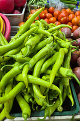A close-up of vibrant green chili peppers displayed at a local farmer's market surrounded by fruits and vegetables.