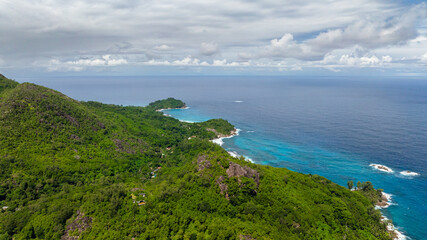 Aerial view of a lush green coastline with hills meeting the blue ocean under a cloudy sky. Seychelles, Mahe.