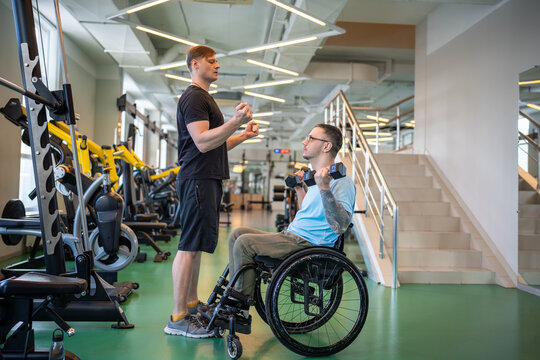 Trainer assists young disabled man in wheelchair lifting dumbbells on personal gym session demonstrating exercise form. Adaptive strength training, spinal injury rehabilitation, physical empowerment