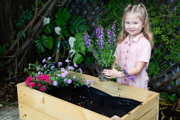 Happy child holding flower pot in backyard – little girl gardening in raised planter box, smiling in front of wooden trellis and green vines, nature education and spring lifestyle concept.